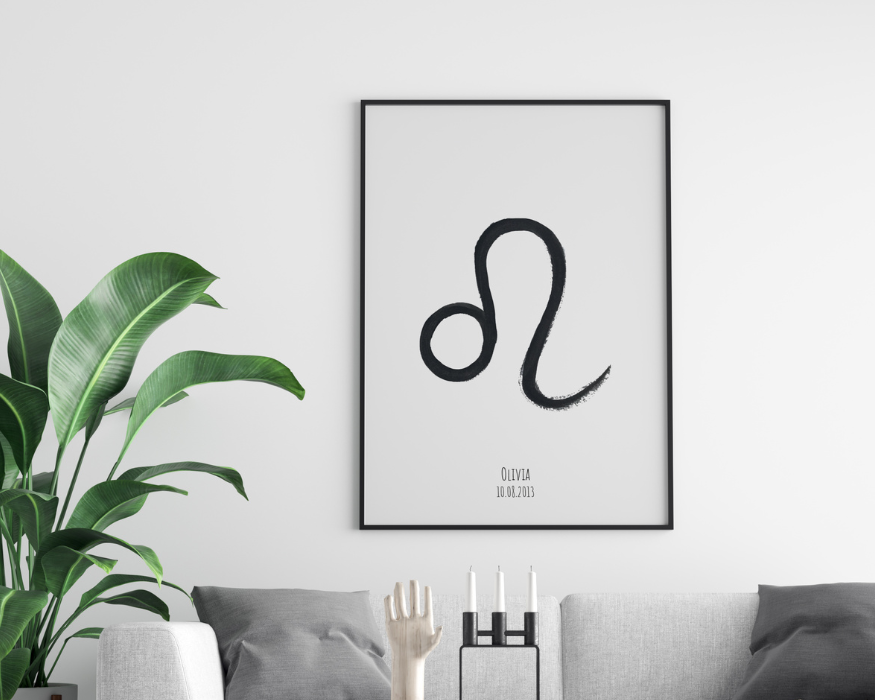 Een ingelijste poster hangend aan een helderwitte muur, direct boven een lichtgrijze bank met donkergrijze kussens. In de strakke zwarte lijst zit een minimalistische, lichtgrijze poster. In het midden staat een groot, met zwarte verf geschilderd astrologisch symbool van het sterrenbeeld Leeuw. Onderaan de poster staat de gepersonaliseerde tekst 'OLIVIA' met daaronder de datum '10.08.2013'. Links van de bank staat een grote, opvallende groene kamerplant met grote bladeren. Voor de poster staan decoratieve items: een houten hand en een moderne zwarte kandelaar met drie witte kaarsen.