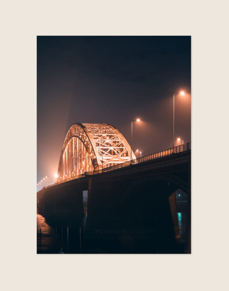 Een sfeervolle nachtfoto van een grote boogbrug, waarschijnlijk de Waalbrug in Nijmegen. De imposante stalen boogconstructie is prachtig verlicht met warm oranje licht. Ook de straatlantaarns op het brugdek stralen een warme gloed uit, wat een mooi contrast vormt met de donkere nachtlucht en de massieve, donkere brugpijlers. De verlichting creëert een lichte waas in de lucht, wat bijdraagt aan het sfeervolle en dramatische beeld.