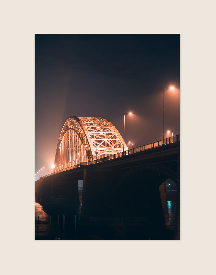 Een sfeervolle nachtfoto van een grote boogbrug, waarschijnlijk de Waalbrug in Nijmegen. De imposante stalen boogconstructie is prachtig verlicht met warm oranje licht. Ook de straatlantaarns op het brugdek stralen een warme gloed uit, wat een mooi contrast vormt met de donkere nachtlucht en de massieve, donkere brugpijlers. De verlichting creëert een lichte waas in de lucht, wat bijdraagt aan het sfeervolle en dramatische beeld.