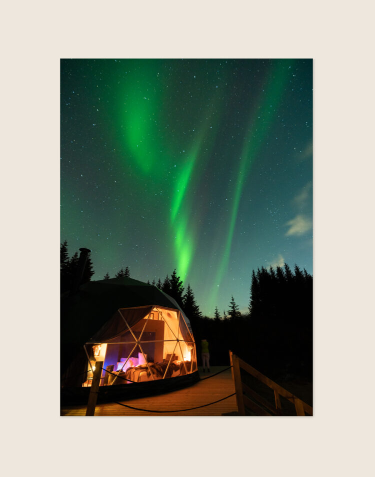 Een spectaculaire nachtfoto van felgroen noorderlicht boven een knusse, warm verlichte glamping dome met een houten vlonder en donkere dennenbomen in IJsland.