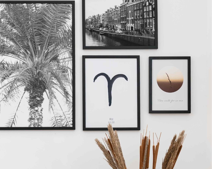 Een sfeervolle fotowand met meerdere posters in strakke zwarte lijsten, hangend op een witte muur. Centraal onderin hangt een gepersonaliseerde poster met een groot, in zwarte verf geschilderd astrologisch symbool van het sterrenbeeld Ram. Daaronder staat de naam 'MIKE' en een datum ('06.04.2005'). De rest van de collage bestaat uit een grote zwart-wit poster van een palmboom, een zwart-wit foto van Amsterdamse grachtenpanden, en een poster met een ronde grafische weergave van een zonsondergang met een grassprietje en de cursieve tekst 'Time waits for no one'. Op de voorgrond steken pluimen van gedroogd gras en rietsigaren in beeld.