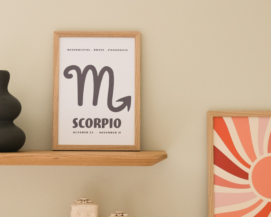 Een ingelijste poster, staand op een houten wandplank tegen een zachte beige muur. In de licht houten lijst zit een lichte poster met bovenaan in kleine letters de tekst 'RESOURCEFUL . BRAVE . PASSIONATE', in het midden een groot donkergrijs astrologisch symbool van de Schorpioen (een 'm' met een pijlpunt aan de staart), en onderaan in dikke, afgeronde letters de tekst 'SCORPIO' met de datum 'OCTOBER 23 - NOVEMBER 21'. Links op de plank staat een stukje van een bobbelige, matzwarte vaas. Rechts in beeld is nog net een deel van een andere ingelijste poster zichtbaar met een retro zonnestralen-patroon in warme roze, oranje en rode tinten.