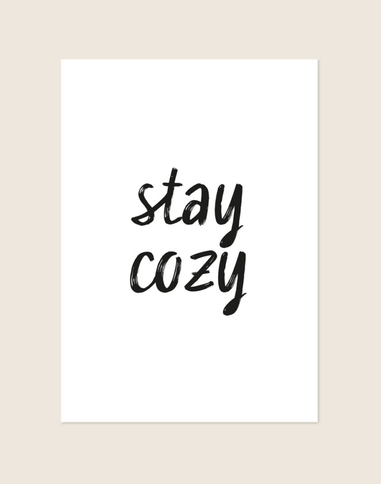 Witte poster met de tekst 'stay cozy' in een zwart, speels en handgeschreven penseel-lettertype, sfeervol opgehangen in een knusse slaapkamer.