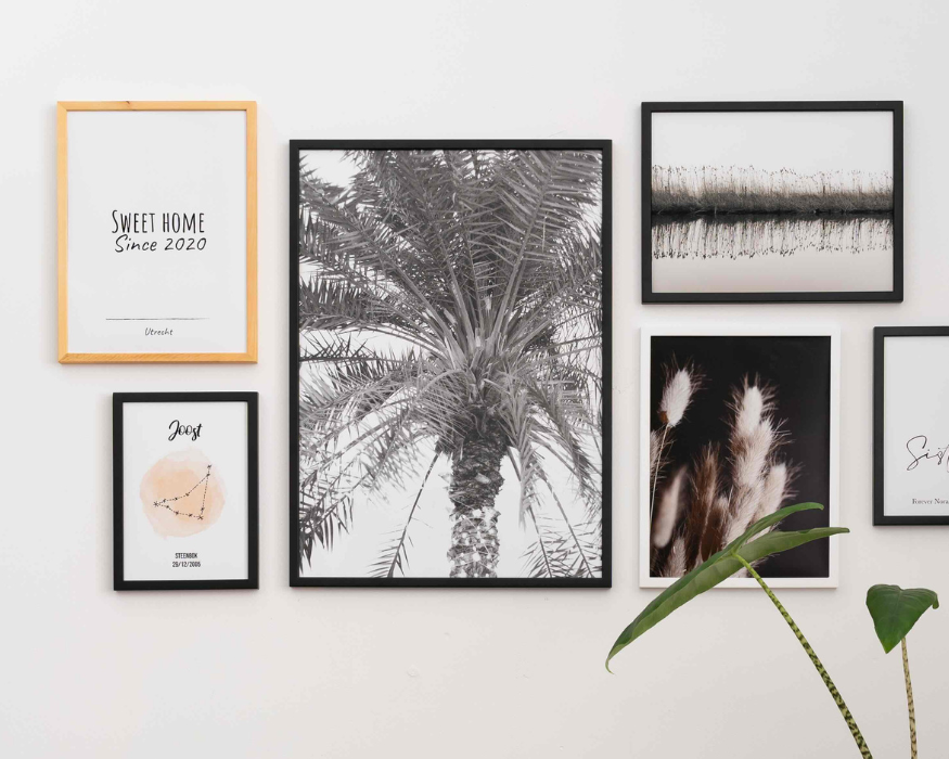 Een sfeervolle fotowand met verschillende ingelijste posters op een helderwitte muur. Linksonder hangt een kleine gepersonaliseerde sterrenbeeld poster in een zwarte lijst met de naam 'Joost', een zachtoranje aquarel vlek met de sterrenconstellatie van de Steenbok en de datum '26/12/2005'. De rest van de collage bestaat uit een poster met de tekst 'SWEET HOME Since 2020 Utrecht', een grote zwart-wit poster van een palmboom, een landschapsfoto van riet langs het water en een donkere poster met lichte pluimgrassen. Rechtsonder is een deel van een opvallende groene kamerplant met een gestreepte stengel zichtbaar.