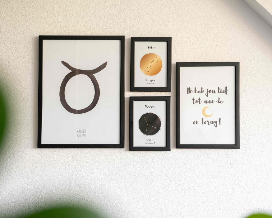 Een fotowand met vier posters in strakke zwarte lijsten, hangend aan een witte, licht getextureerde muur. Aan de linkerkant hangt een grote gepersonaliseerde poster met een groot, in zwarte verf geschilderd astrologisch symbool van het sterrenbeeld Stier, met daaronder de naam 'MAARTJE' en de datum '26.04.1989'. In het midden hangen twee kleinere posters boven elkaar: de bovenste heeft een oranje vlek met het sterrenbeeld Schorpioen, de naam 'Fien' en een datum, de onderste een zwarte vlek met het sterrenbeeld Kreeft, de naam 'Bram' en een datum. Aan de rechterkant hangt een middelgrote poster met de tekst 'Ik heb jou lief tot aan de [illustratie van een gouden maantje] en terug!'. Op de wazige voorgrond zijn groene bladeren van een kamerplant zichtbaar.