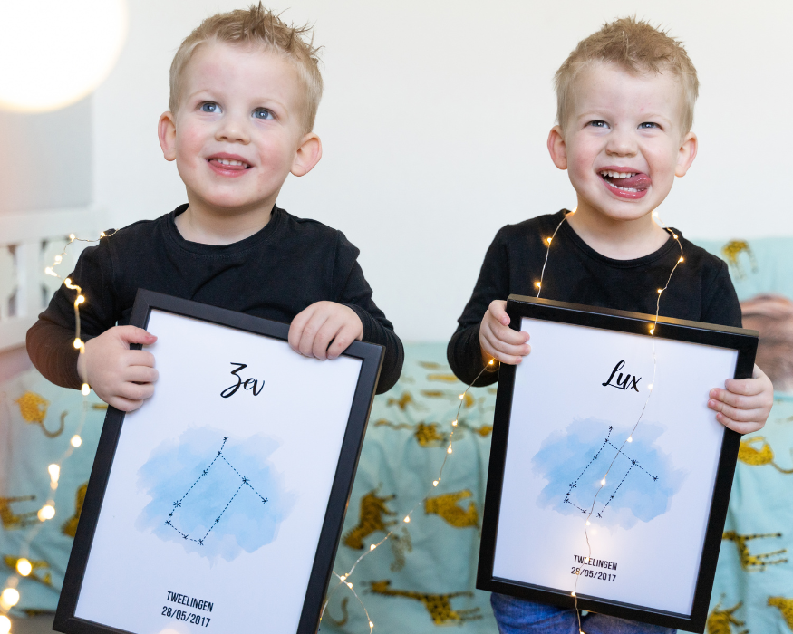 Een foto van twee jonge tweelingbroertjes in zwarte shirts die elk een ingelijste gepersonaliseerde sterrenbeeld-poster vasthouden in een sfeervol verlichte kinderkamer. De zwarte fotolijsten zijn omhuld door brandende sfeerverlichting (fairy lights). De linker poster is gepersonaliseerd met de naam 'Zev' in sierlijke letters, een blauwe waterverfvlek met het verbonden Gemini-sterrenbeeld, de tekst 'TWEELINGEN' en de datum '28/05/2017'. De rechter poster is identiek, maar met de naam 'Lux'. De jongen aan de rechterkant steekt zijn tong uit en kijkt recht in de camera. Ze staan voor een bed met beddengoed met een safaripatroon.