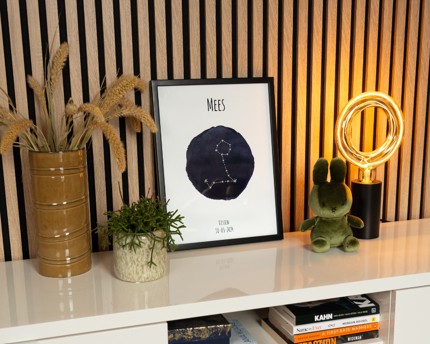 Een ingelijste gepersonaliseerde poster, staand op een glanzend wit meubel tegen een achterwand met verticale houten latten. In de strakke zwarte lijst zit een poster met de naam 'MEES', een donkerblauwe aquarel vlek met daarin de witte sterrenconstellatie van Vissen, en onderaan de tekst 'VISSEN' met de datum '10-03-2024'. Rondom de poster staan sfeervolle woonaccessoires: links een bruine vaas met gedroogde grassen en een klein groen plantje in een gespikkelde pot, en rechts een donkergroene fluwelen Nijntje-knuffel naast een moderne zwarte tafellamp met een opvallende grote, ronde lichtbron. In het open vak onder de poster liggen diverse boeken opgestapeld.