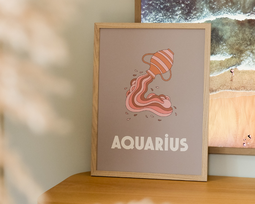 Een ingelijste poster, staand op een houten meubel. In de licht houten lijst zit een poster met een zachte, taupekleurige achtergrond en een retro-stijl illustratie van een oranje met roze gestreepte kruik waaruit golvende stromen water vloeien. Onderaan de poster staat in opvallende, dikke witte letters het woord 'AQUARIUS'. Op de achtergrond staat een grotere lijst met een foto van een strandtafereel en links op de wazige voorgrond is een lichte pluim van gedroogd gras of bloemen zichtbaar.