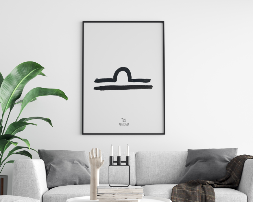 Een ingelijste poster hangend aan een helderwitte muur, direct boven een lichtgrijze bank met donkergrijze kussens en een bruin geruite deken. In de strakke zwarte lijst zit een minimalistische, lichtgrijze poster. In het midden staat een groot, met zwarte verf geschilderd astrologisch symbool van het sterrenbeeld Weegschaal. Onderaan de poster staat de gepersonaliseerde tekst 'Tess' met daaronder de datum '28.09.2002'. Links van de bank staat een grote, opvallende groene kamerplant. Op de voorgrond staan decoratieve items op een stapel boeken: een houten hand en een moderne zwarte kandelaar met drie witte kaarsen.