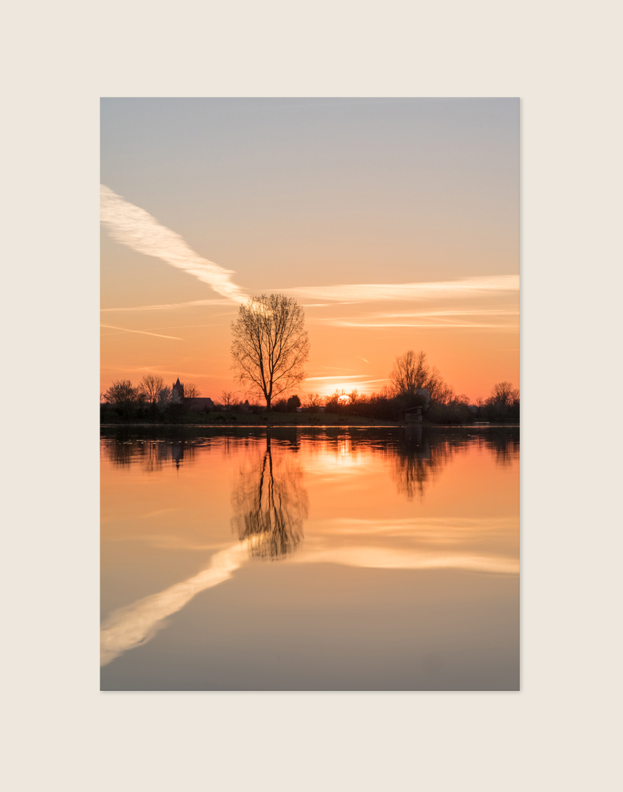 Een landschapsfoto van een zonsondergang boven een glad wateroppervlak. Aan de horizon staan donkere silhouetten van kale bomen en een kerktoren. De oranje lucht, de laagstaande zon en een diagonale condensstreep weerspiegelen symmetrisch in het water op de voorgrond.