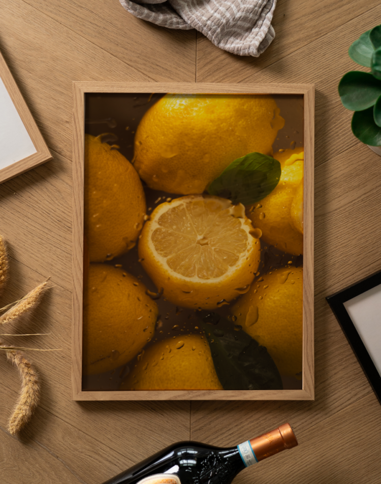 Een sfeervolle flatlay foto (van bovenaf genomen) op een lichtbruine houten vloer of tafel met een visgraatmotief. Centraal ligt een poster in een lichte houten lijst. De poster toont een donkere, sfeervolle close-up foto van heldergele citroenen, rijkelijk bedekt met waterdruppels. In het midden ligt een prachtig doorgesneden halve citroen, speels afgewisseld met enkele donkergroene blaadjes. Rondom de fotolijst liggen diverse decoratieve objecten: bovenaan een opgevouwen beige theedoek met witte streepjes, links enkele gedroogde graanhalmen en het hoekje van een andere lichte houten fotolijst. Rechtsboven is een stukje van een groen kamerplantje zichtbaar, rechtsonder de hoek van een zwarte fotolijst, en onderaan ligt de hals van een donkere wijnfles met een koperkleurige dop.
