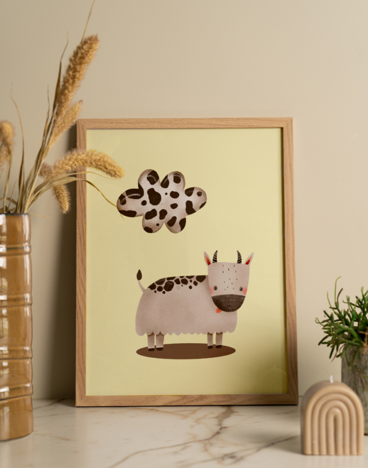 Een sfeervolle interieuropstelling met een ingelijste poster, staand op een licht marmeren oppervlak met subtiele donkere aderen, tegen een zachtbeige muur. In de lichte houten lijst zit een schattige kinderkamer poster met een zachtgele achtergrond. Op de poster staat een lieve, gestileerde illustratie van een bolle, lichtgrijze koe met donkere vlekken op haar rug, kleine hoorntjes, roze wangetjes en een schattig uitstekend rood tongetje. Boven de koe zweeft een speelse wolk die ook is voorzien van een zwart-wit koeienvlekkenpatroon. Aan de linkerkant staat een hoge, geribbelde okergele glanzende vaas met daarin pluizige gedroogde grassen. Aan de rechterkant staat een klein, groen sprieterig plantje in een licht gespikkelde pot, met op de voorgrond een decoratieve beige, boogvormige ribbelkaars.
