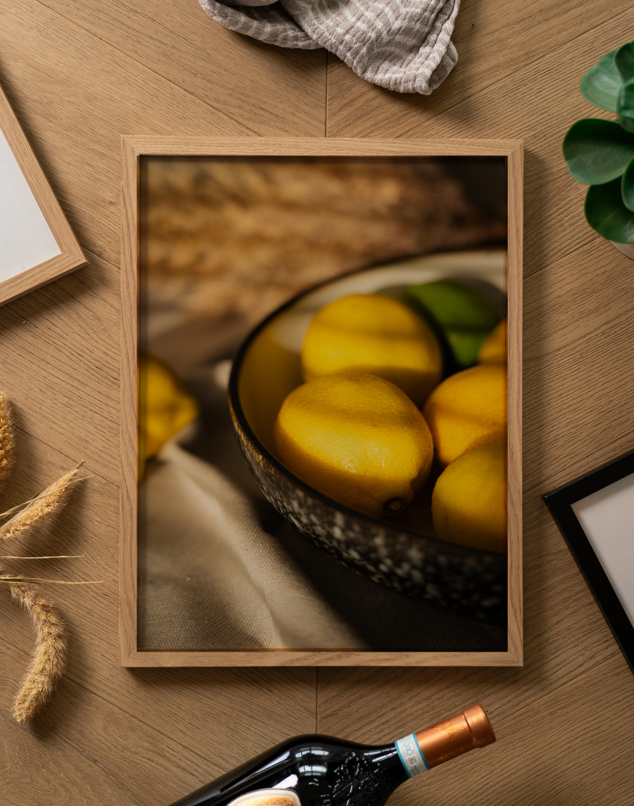 Een sfeervolle flatlay foto (van bovenaf genomen) op een lichtbruine houten ondergrond met een visgraatmotief. Centraal ligt een poster in een lichte houten lijst. De poster toont een warme, sfeervolle stilleven-foto van een donkere, gespikkelde schaal gevuld met heldergele citroenen, waar prachtig licht en schaduw overheen valt. De schaal staat op een soepel vallende, gedrapeerde lichte doek met op de achtergrond onscherpe accenten van gedroogde grassen. Rondom de fotolijst liggen diverse decoratieve objecten: bovenaan een opgevouwen gestreepte beige theedoek, links enkele gedroogde graanhalmen en het hoekje van een andere lichte houten fotolijst. Rechtsboven is een stukje van een groen kamerplantje zichtbaar, rechtsonder de hoek van een strakke zwarte fotolijst, en onderaan de hals van een donkere wijnfles met een koperkleurige capsule.