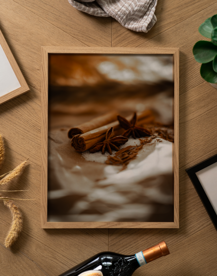 Een sfeervolle flatlay foto (van bovenaf genomen) op een lichtbruine houten ondergrond met een visgraatmotief. Centraal ligt een poster in een lichte houten lijst. De poster toont een warme, sfeervolle close-up foto (macro) van winterse specerijen. In scherpe focus liggen enkele kaneelstokjes en stervormige steranijs op een bedje van witte suikerkristallen en gestrooide kruiden, rustend op gekreukeld papier. De voor- en achtergrond hebben een zachte, dromerige waas (bokeh). Rondom de fotolijst liggen diverse decoratieve objecten: bovenaan een opgevouwen gestreepte beige theedoek, links enkele gedroogde graanhalmen en het hoekje van een andere lichte houten fotolijst. Rechtsboven is een stukje van een groen kamerplantje zichtbaar, rechtsonder de hoek van een strakke zwarte fotolijst, en onderaan de hals van een donkere wijnfles met een koperkleurige capsule.