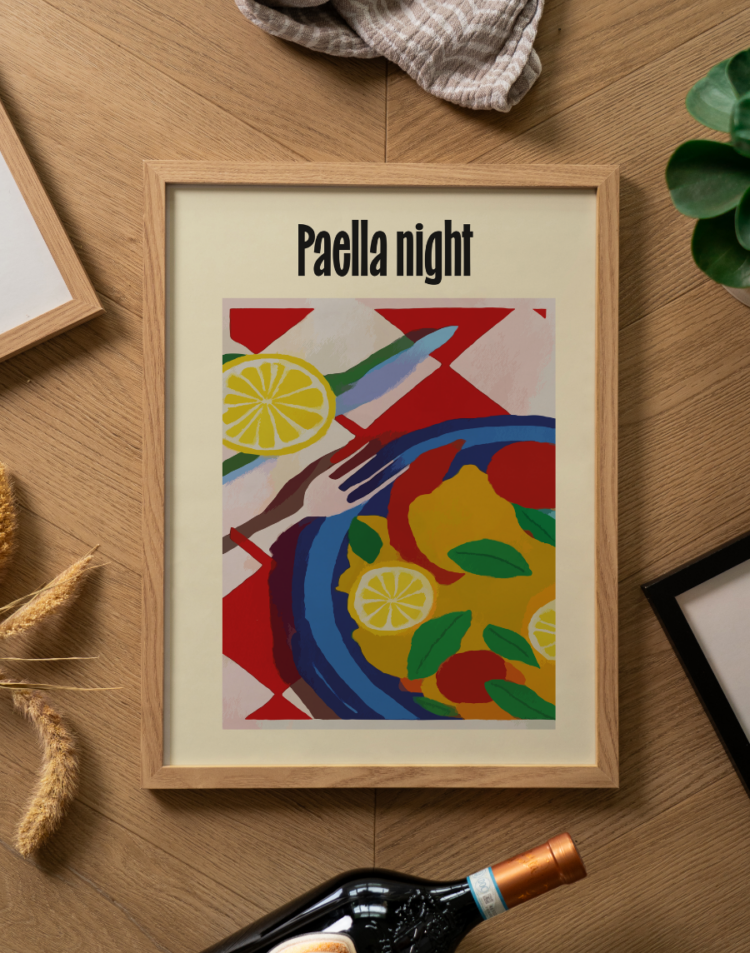 Een sfeervolle flatlay foto (van bovenaf genomen) op een lichtbruine houten ondergrond met een visgraatmotief. Centraal ligt een poster in een lichte houten lijst. De poster heeft een crèmekleurige achtergrond met bovenaan in opvallende, zwarte letters de tekst 'Paella night'. Daaronder staat een kleurrijke, abstract geschilderde illustratie van een paellamaaltijd. Je ziet een donkerblauwe pan op een rood-wit geblokt tafelkleed, gevuld met gele rijst, rode en groene accenten, en felgele schijfjes citroen. Naast de pan liggen een gestileerde vork en een mes. Rondom de fotolijst liggen diverse decoratieve objecten: bovenaan een opgevouwen gestreepte beige theedoek, links enkele gedroogde graanhalmen en het hoekje van een andere lichte houten fotolijst. Rechtsboven is een stukje van een groen kamerplantje zichtbaar, rechtsonder de hoek van een strakke zwarte fotolijst, en onderaan de hals van een donkere wijnfles met een koperkleurige capsule.