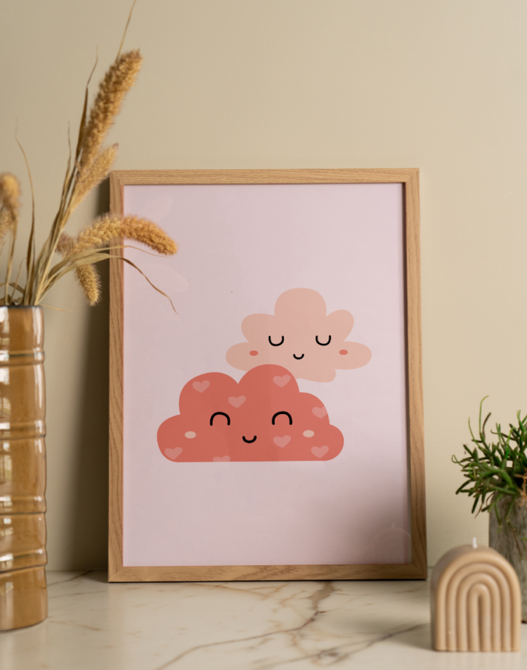 Een sfeervolle interieuropstelling met een ingelijste poster, staand op een licht marmeren oppervlak met subtiele donkere aderen, tegen een zachtbeige muur. In de lichte houten lijst zit een schattige kinderkamer poster met een zachtroze achtergrond. Centraal op de poster staan twee gestileerde, vrolijke wolkjes met schattige gezichtjes (gesloten boogvormige oogjes en een glimlachje). De onderste, grotere wolk is donkerroze gekleurd en versierd met kleine, lichtroze hartjes. Het bovenste, kleinere wolkje is effen zachtroze. Aan de linkerkant staat een hoge, geribbelde okergele glanzende vaas met daarin pluizige gedroogde grassen. Aan de rechterkant staat een klein, groen sprieterig plantje in een licht gespikkelde pot, met op de voorgrond een decoratieve beige, boogvormige ribbelkaars.