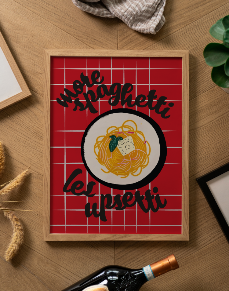 Een sfeervolle flatlay foto (van bovenaf genomen) op een lichtbruine houten ondergrond met een visgraatmotief. Centraal ligt een poster in een lichte houten lijst. De opvallende poster heeft een felrode achtergrond met een wit ruitpatroon, vergelijkbaar met een klassiek Italiaans tafelkleed. Bovenaan staat in dikke, speelse zwarte letters de tekst 'more spaghetti' en onderaan maakt de tekst 'less upsetti' de humoristische rijm compleet. In het midden staat een grafische illustratie van een wit bord met een zwarte rand, gevuld met een nestje gele spaghetti, afgetopt met een klontje boter met zwarte peper en een groen basilicumblaadje. Rondom de fotolijst liggen diverse decoratieve objecten: bovenaan een opgevouwen gestreepte beige theedoek, links enkele gedroogde graanhalmen en het hoekje van een andere lichte houten fotolijst. Rechtsboven is een stukje van een groen kamerplantje zichtbaar, rechtsonder de hoek van een strakke zwarte fotolijst, en onderaan de hals van een donkere wijnfles met een koperkleurige capsule.