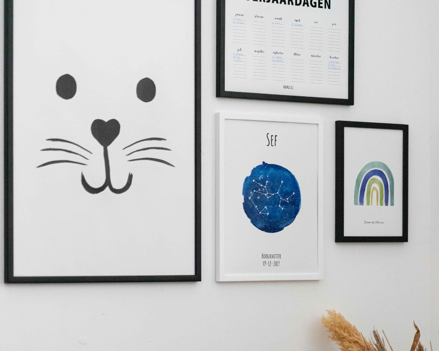 Een sfeervolle collage van ingelijste posters op een witte muur. Centraal onderin hangt een gepersonaliseerde sterrenbeeld poster in een witte lijst met de naam 'SEF', een diepblauwe aquarel vlek met daarin de sterrenconstellatie van de Boogschutter, en onderaan de tekst 'BOOGSCHUTTER' met de datum '09-12-2019'. Verder bestaat de fotowand uit een grote poster in een zwarte lijst met een minimalistisch zwart-wit dierengezichtje (met een hartje als neus), een verjaardagskalender poster ('VERJAARDAGEN') en een kleine poster met een blauw-groene aquarel regenboog en de tekst 'Dream big little one'.