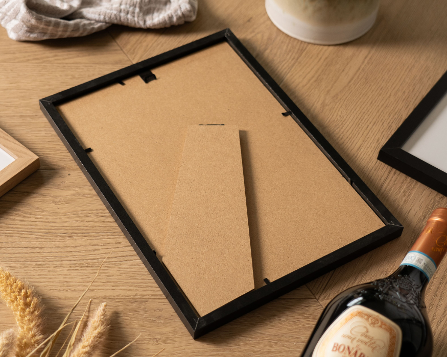De achterkant van een zwarte fotolijst, diagonaal liggend op een houten visgraatvloer of -tafel. De achterkant is gemaakt van bruin hardboard en is voorzien van een uitklapbare standaard en zwarte sluitklemmetjes. Rondom de lijst liggen sfeervolle details, waaronder een donkere glazen wijnfles met een gouden hals, gedroogde grassen, een opgevouwen linnen doek en de hoeken van twee andere fotolijstjes.