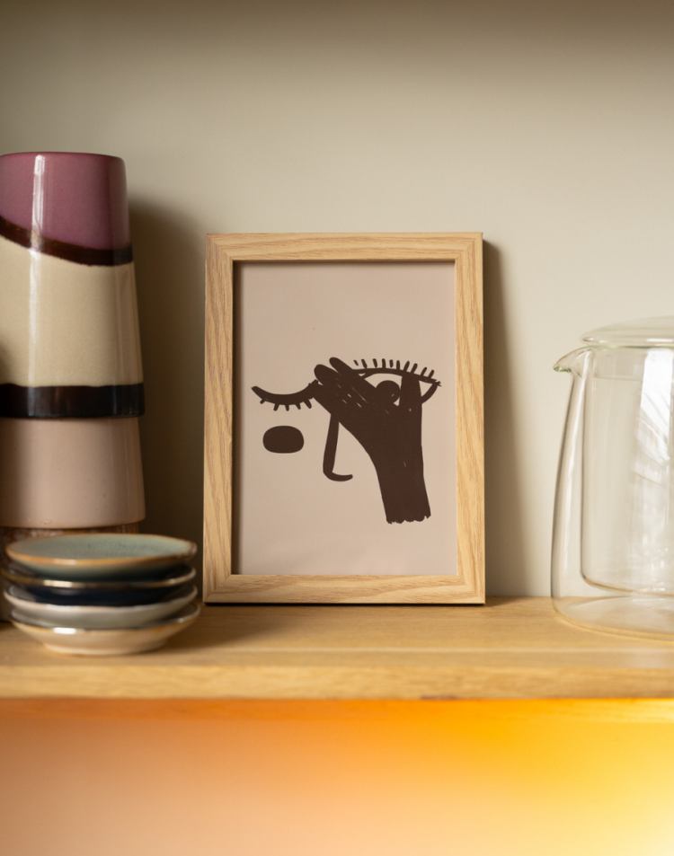 Een ingelijste poster op een houten plank. De afbeelding toont een beige achtergrond met een donkerbruine, abstracte illustratie van een hand die voor een oog wordt gehouden. Links van de lijst staan gestapelde, meerkleurige keramieken bekers en rechts staat een glazen kan.