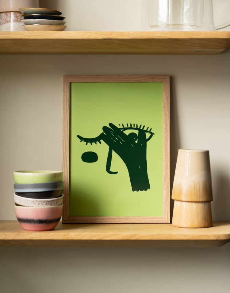 Een ingelijste poster op een houten plank. Deze variant heeft een felgroene achtergrond met een donkergroene, abstracte illustratie van een hand voor een oog. Links staan kleurrijke gestapelde schaaltjes en rechts staat een tweekleurige keramieken vaas.