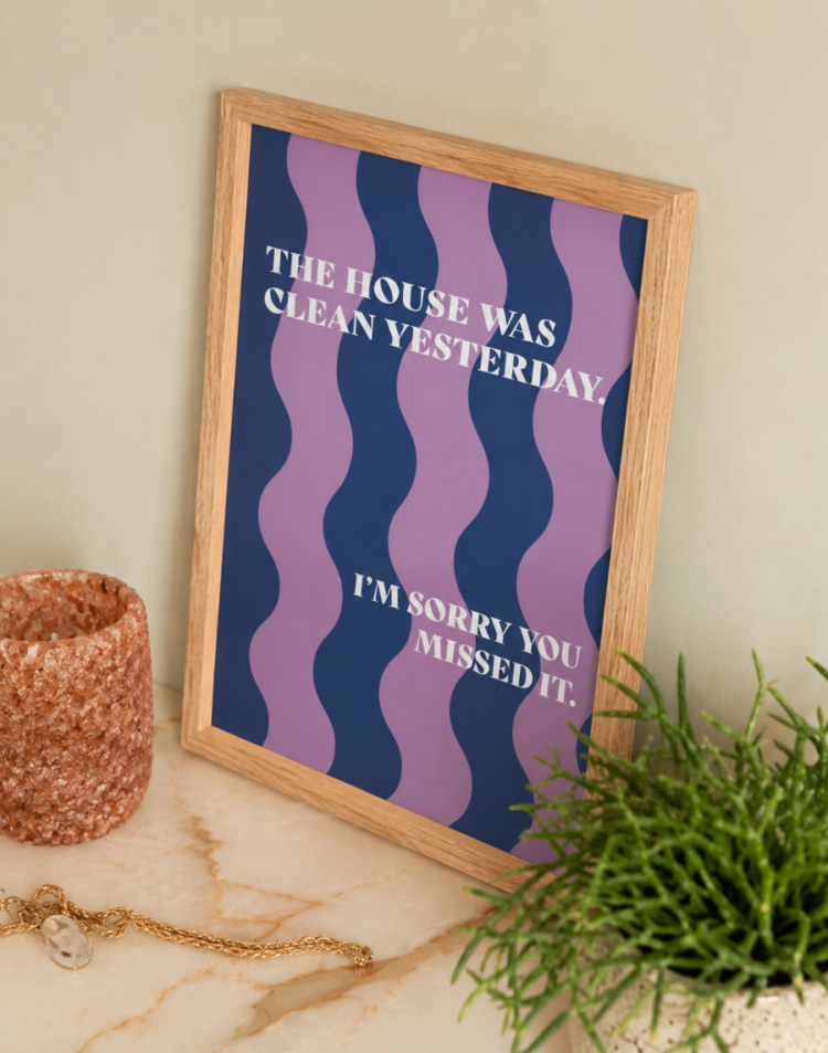 Een ingelijste poster met een humoristische tekst staat op een marmeren oppervlak. De poster heeft een patroon van golvende paarse en donkerblauwe strepen en de witte tekst luidt: "THE HOUSE WAS CLEAN YESTERDAY. I'M SORRY YOU MISSED IT.". Er staan een kleine potplant en een getextureerde vaas naast de lijst.