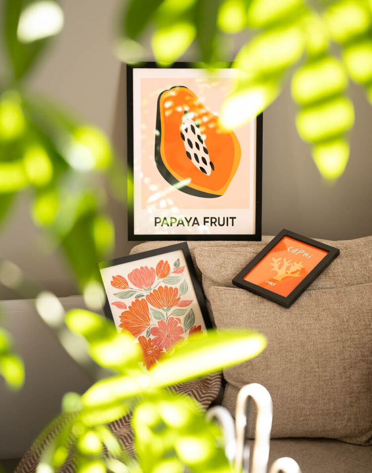 Drie ingelijste posters op en achter een beige bank. Bovenaan staat een poster van een opengesneden papaja met de tekst 'PAPAYA FRUIT'. Linksonder ligt een lijst met oranje en roze bloemen, en rechts een kleine lijst met een oranje illustratie en de tekst 'CAPRI ITALY'. Op de voorgrond zijn wazige groen-gele bladeren te zien.