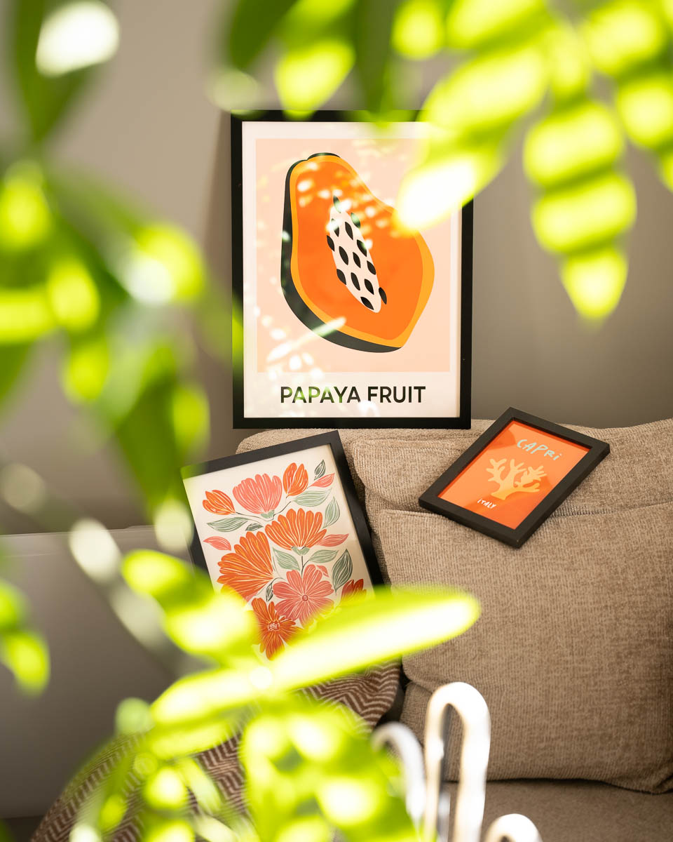Drie ingelijste posters op en achter een beige bank. Bovenaan staat een poster van een opengesneden papaja met de tekst 'PAPAYA FRUIT'. Linksonder ligt een lijst met oranje en roze bloemen, en rechts een kleine lijst met een oranje illustratie en de tekst 'CAPRI ITALY'. Op de voorgrond zijn wazige groen-gele bladeren te zien.