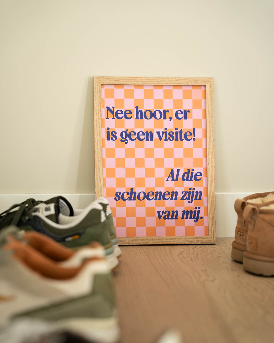 Een houten schilderijlijst op een houten vloer tegen een beige muur. De poster heeft een roze-oranje blokkenpatroon met de blauwe tekst: "Nee hoor, er is geen visite! Al die schoenen zijn van mij." Op de voorgrond en aan de zijkant staan verschillende schoenen.