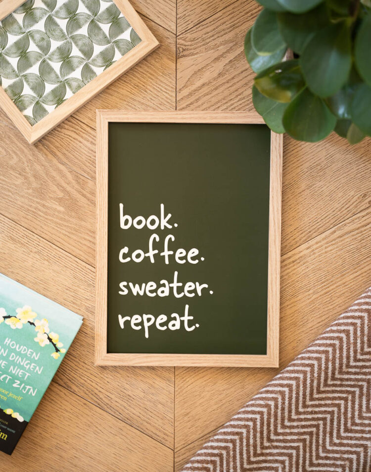 Bovenaanzicht van een donkergroene poster in een lichte houten lijst met de witte tekst "book. coffee. sweater. repeat." De lijst ligt op een houten visgraatvloer, omringd door een kleiner ingelijst groen patroon, een boek, een plant en een deken.