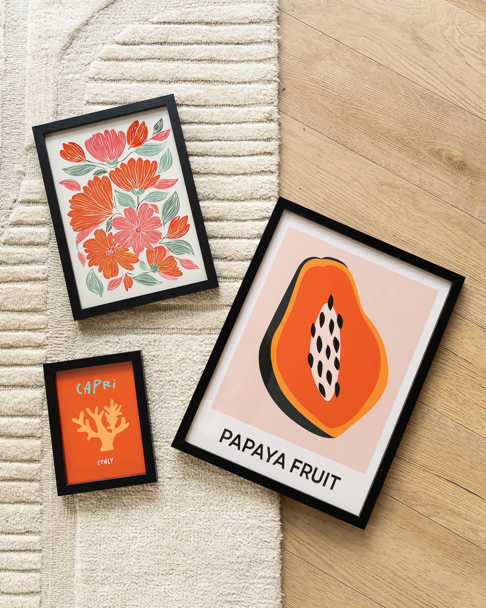 Drie zwarte schilderijlijsten op een licht beige vloerkleed en houten vloer. De gelaagde opstelling bevat een grote poster van een opengesneden papaja, een poster met een drukke illustratie van oranje en roze bloemen, en een kleine oranje poster met een koraalmotief en de tekst 'CAPRI ITALY'.