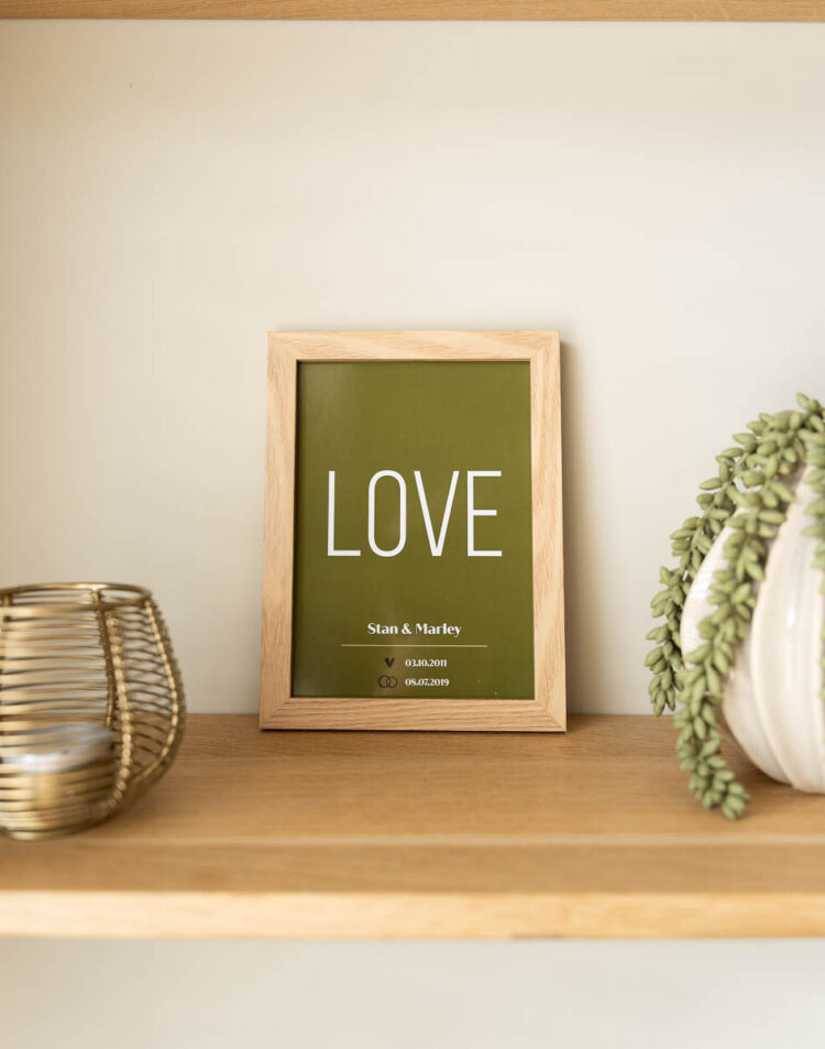 Een olijfgroene poster in een houten lijst op een eikenhouten plank. Op de poster staat het woord 'LOVE' met eronder de namen 'Stan & Marley' en twee data met icoontjes. Rondom de lijst staan een goudkleurige waxinelichthouder en een vaas met een hangplant.