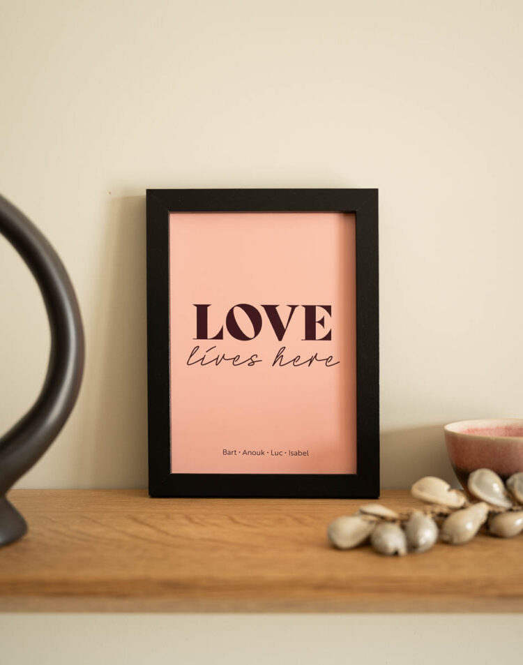 Een roze poster in een strakke zwarte lijst op een houten plank. Op de poster staat de tekst "LOVE lives here" met onderaan de namen "Bart · Anouk · Luc · Isabel". Naast de lijst staan een decoratieve zwarte vorm, een schaaltje en een schelpenketting.