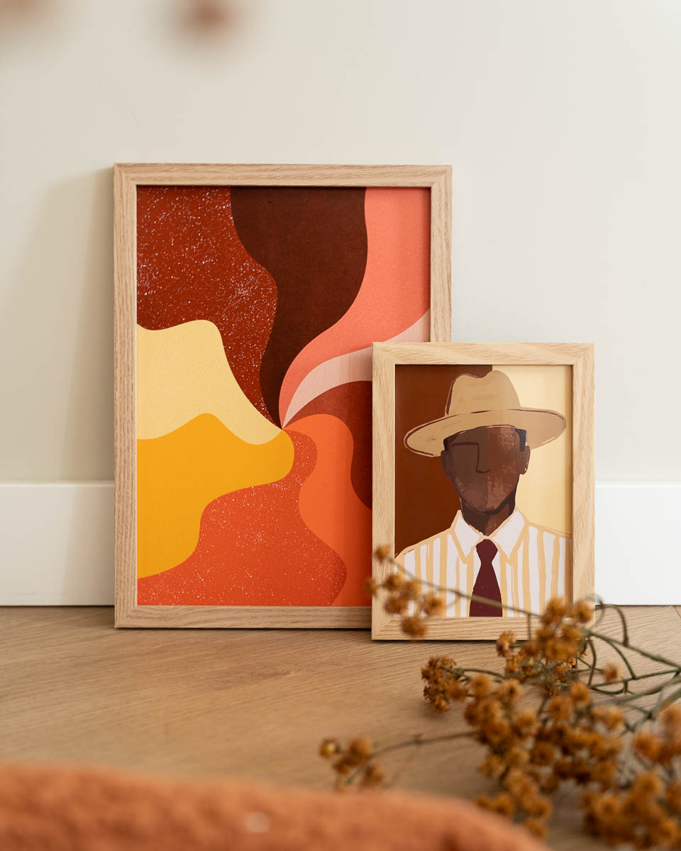 Twee houten schilderijlijsten op een houten vloer tegen een witte muur. Links staat een grote poster met een abstract, golvend patroon in roestbruin, oranje, roze en geel. Rechtsvoor staat een kleinere poster van een man met een hoed. Op de voorgrond liggen gedroogde bloemen.