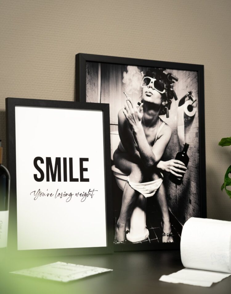 Twee zwarte schilderijlijsten op een donkere ondergrond. De voorste poster heeft een witte achtergrond met de tekst "SMILE You're losing weight". De achterste poster is een zwart-witfoto van een vrouw met zonnebril en krulspelden die op het toilet zit met een fles wijn. Rechtsvoor ligt een rol toiletpapier.