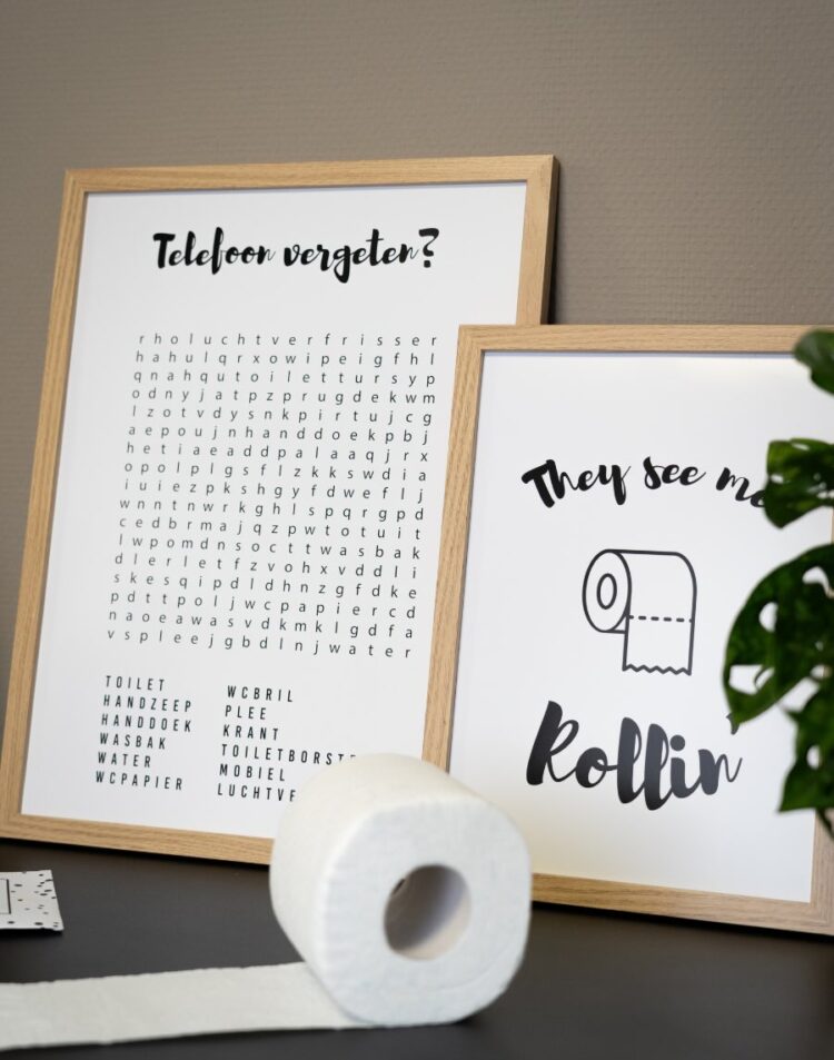 Twee posters in lichte houten lijsten. De linker poster toont een woordzoeker met de titel "Telefoon vergeten?". De rechter poster toont een illustratie van een toiletrol met de tekst "They see me Rollin'". Op de voorgrond staat een rol toiletpapier.