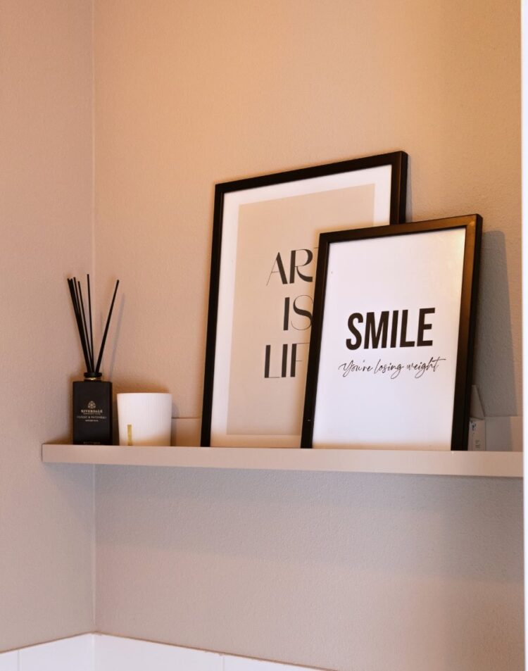 Twee zwarte schilderijlijsten op een wandplank tegen een taupekleurige muur. De voorste poster vermeldt "SMILE You're losing weight" en de achterste toont gedeeltelijk de tekst "ART IS LIFE". Links staan geurstokjes en een klein potje.