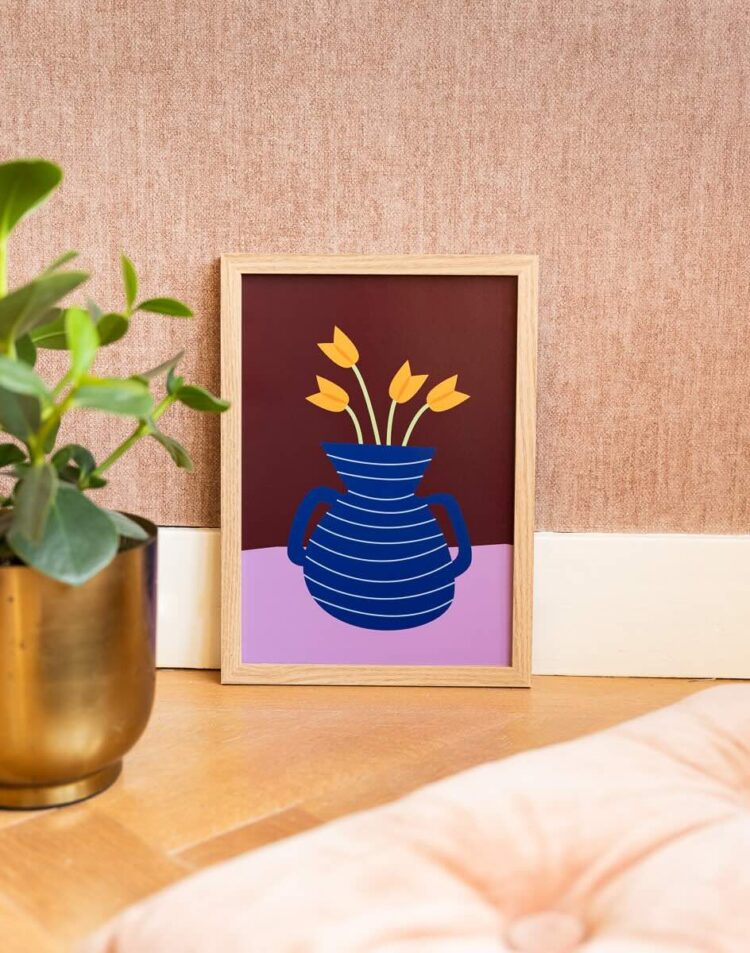 Een houten lijst met een grafische poster staat op de vloer tegen een roze behangen muur. De poster toont een blauwe vaas met gele tulpen tegen een donkerrode en lila achtergrond. Links staat een gouden bloempot met een groene plant en rechtsonder ligt een lichtroze kussen.
