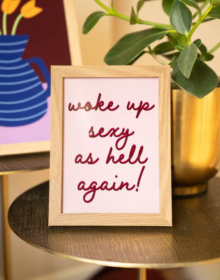 Een ingelijste poster op een gouden bijzettafeltje met de rode geschreven tekst "wake up sexy as hell again!" op een lichtroze achtergrond. Op de achtergrond staat een poster met een blauwe vaas en rechts staat een goudkleurige pot met een groene plant.