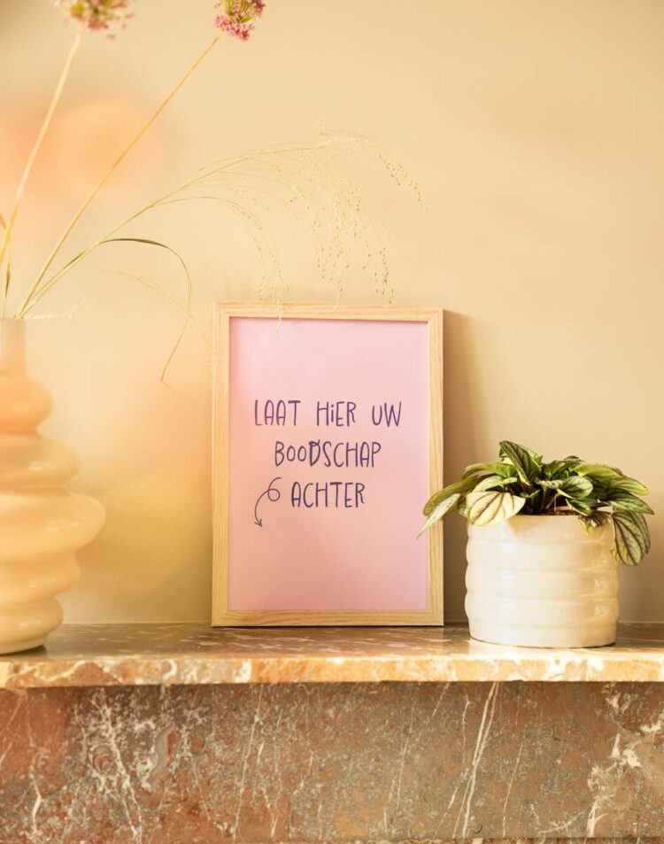 Een roze poster in een houten lijst op een marmeren schouw met de handgeschreven tekst "LAAT HIER UW BOODSCHAP ACHTER" en een pijl. Naast de lijst staat een klein wit potje met een plantje en links staat een vaas met droogbloemen.
