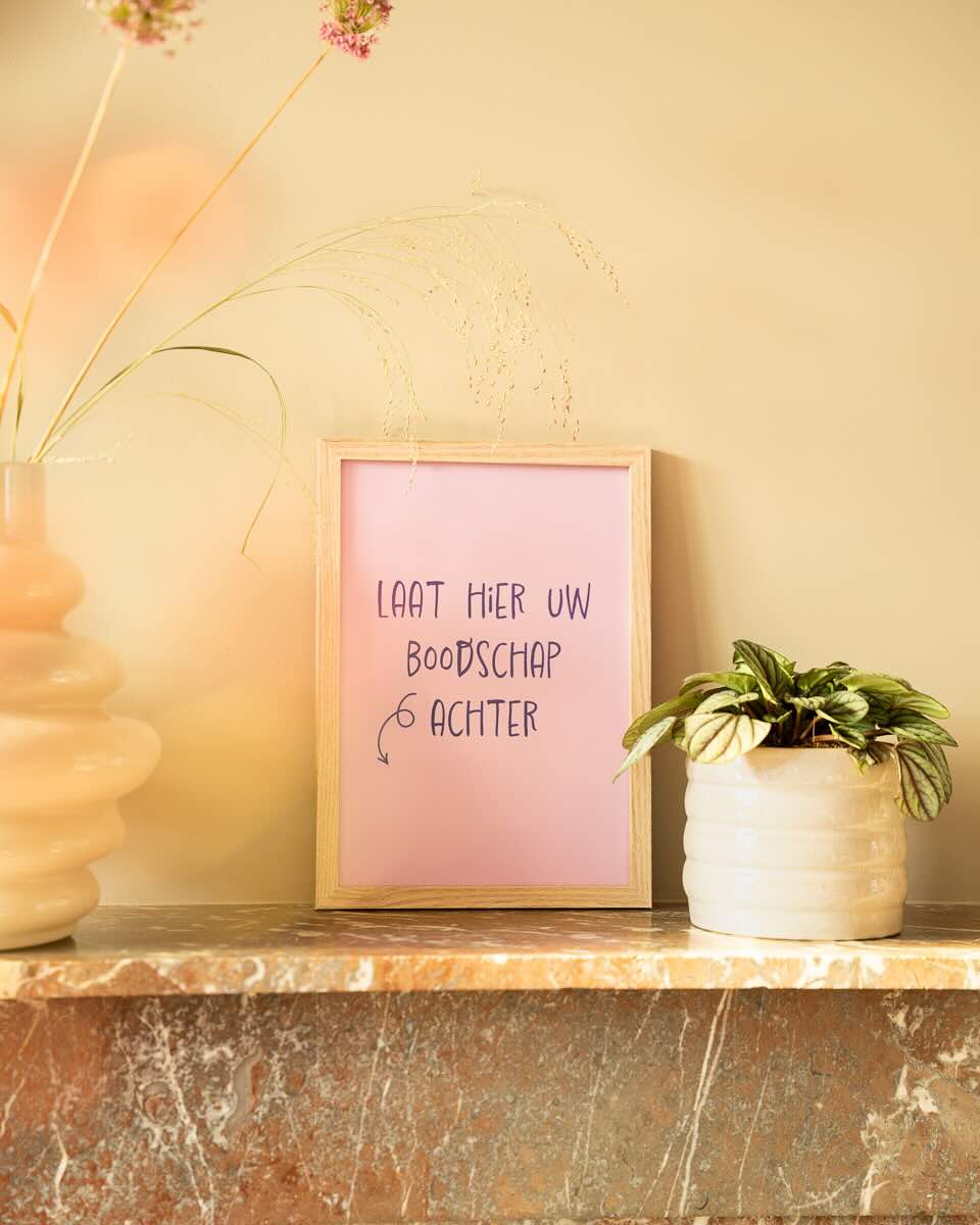 Een roze poster in een houten lijst op een marmeren schouw met de handgeschreven tekst "LAAT HIER UW BOODSCHAP ACHTER" en een pijl. Naast de lijst staat een klein wit potje met een plantje en links staat een vaas met droogbloemen.