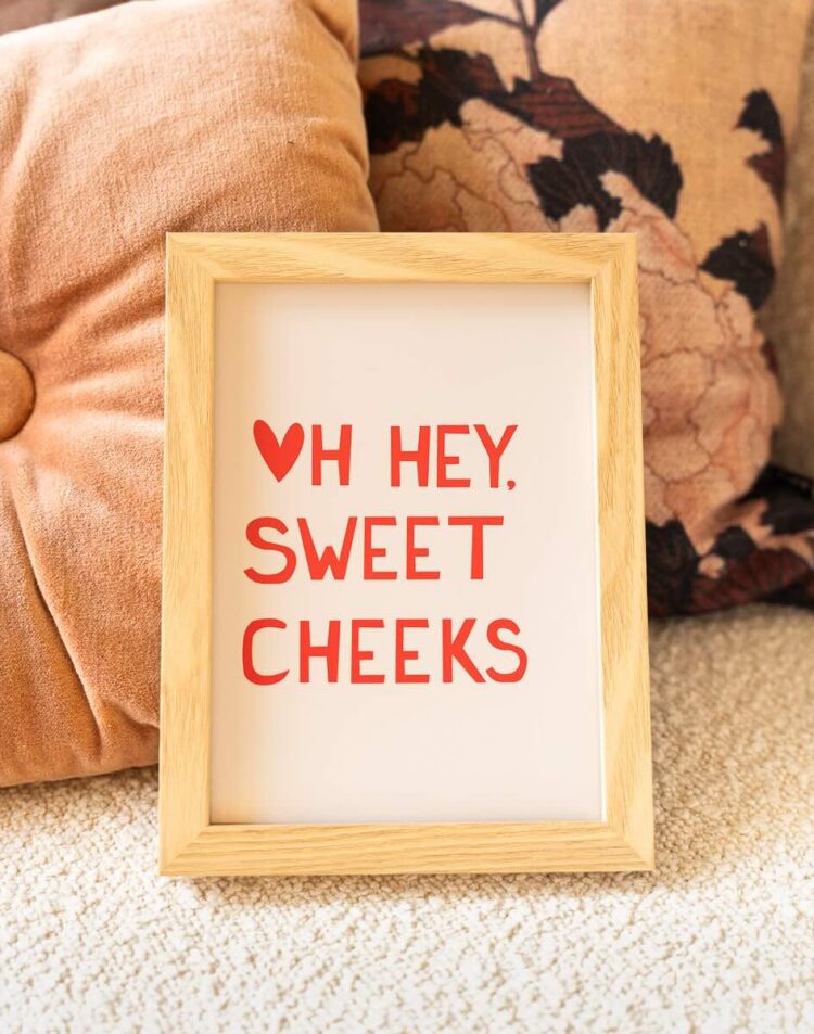 Een ingelijste poster leunt tegen een roze en een drukke bloemen-sierkussen op een zachte ondergrond. Op de poster staat in rode letters de tekst "♥ HEY, SWEET CHEEKS".