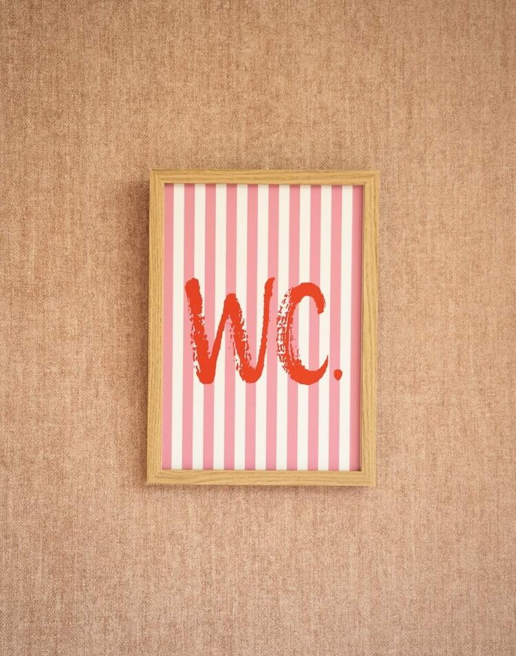 Een ingelijste poster hangt op een roze muur met textuur. De poster heeft een roze met wit verticaal gestreept patroon waarop in rode, geschilderde letters "WC." staat geschreven.